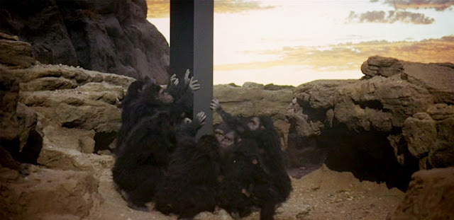 Scene from 2001 with primitive humanoids pawing at monolith.